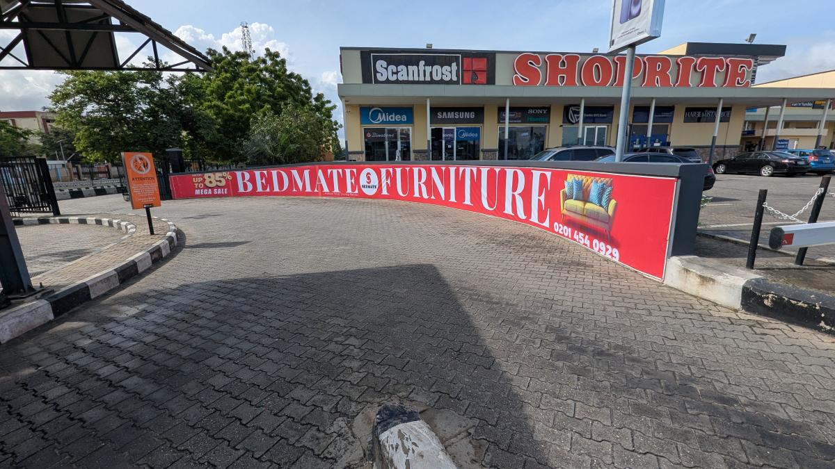 The billboard is strategically positioned along the curved concrete-brick paved driveway at a major commercial hub, the Ikeja City Mall.